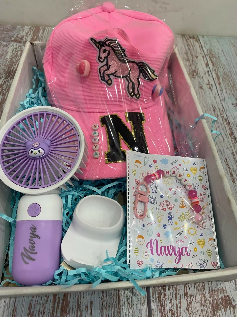 Pink cap with unicorn design, purple fan, white container, and notebook with 'Navya' branding in a box.