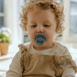 Child wearing a beige outfit with a blue pacifier indoors