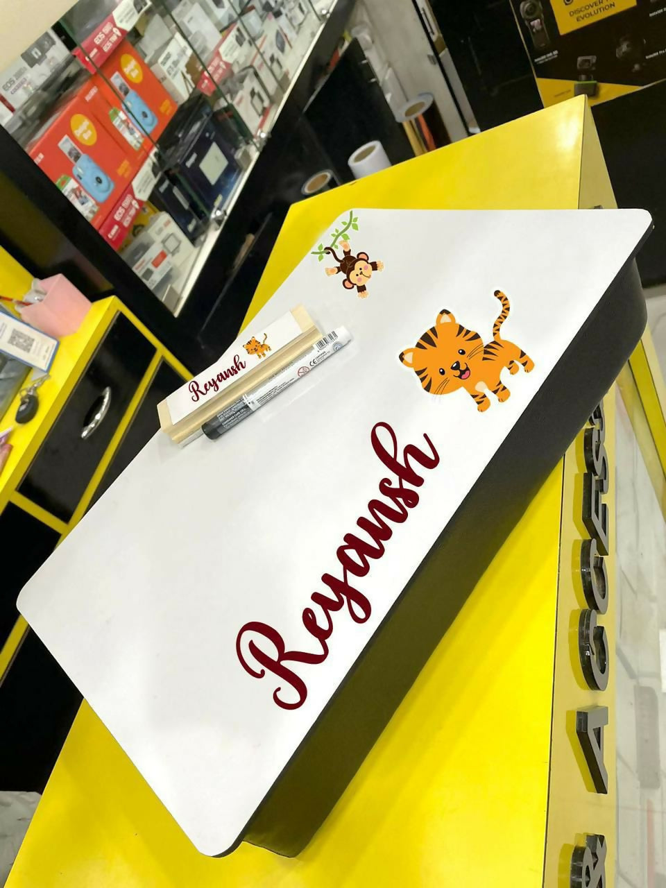 White board with 'Reyansh' written on it, placed on a yellow surface with cartoon characters.