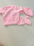 Pink baby sweater, hat, and booties set on a light gray surface