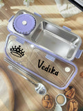 Stainless steel lunch box with purple lid, spoon, and cookies on a wooden surface