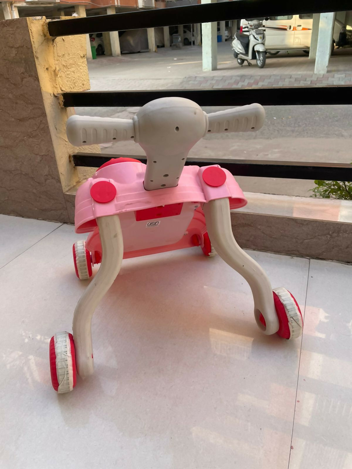 Pink and white children's tricycle on a tiled floor with a scooter in the background.