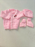 Pink baby cardigan, hat, and booties set on a textured surface