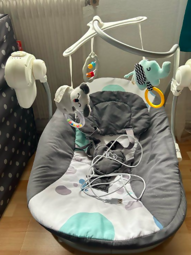 Gray baby bouncer with toys and a white cord on a wooden floor.