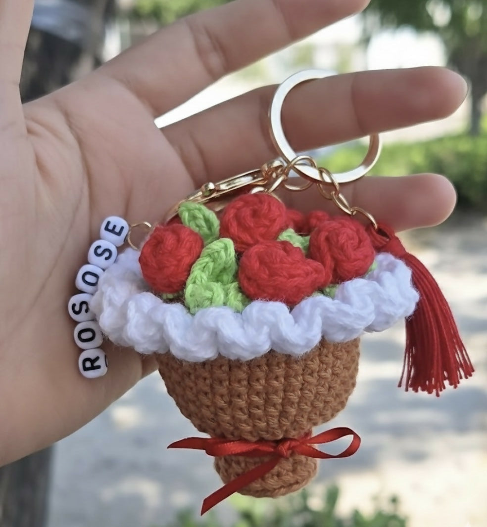 Hand holding a crocheted keychain shaped like a cupcake with red roses, green leaves, and a red bow.
