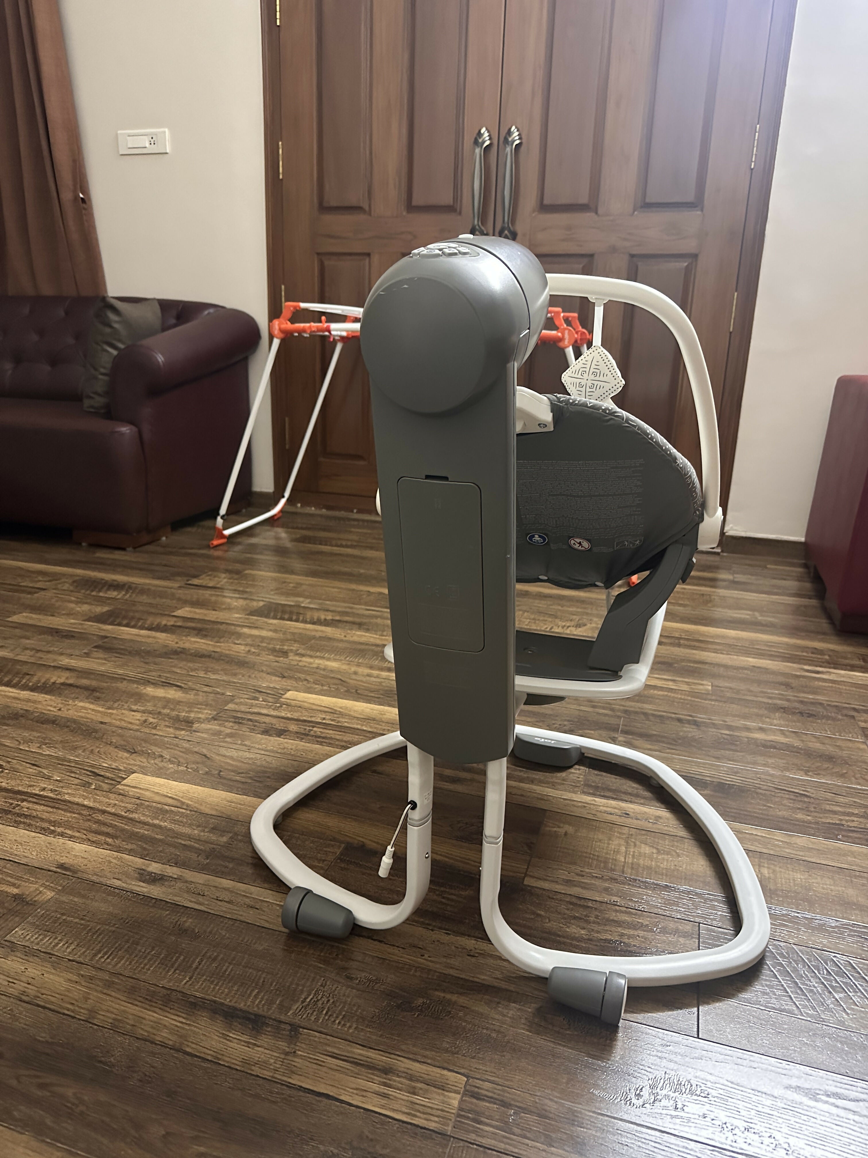 Baby swing on a wooden floor with a brown couch and wooden door in the background