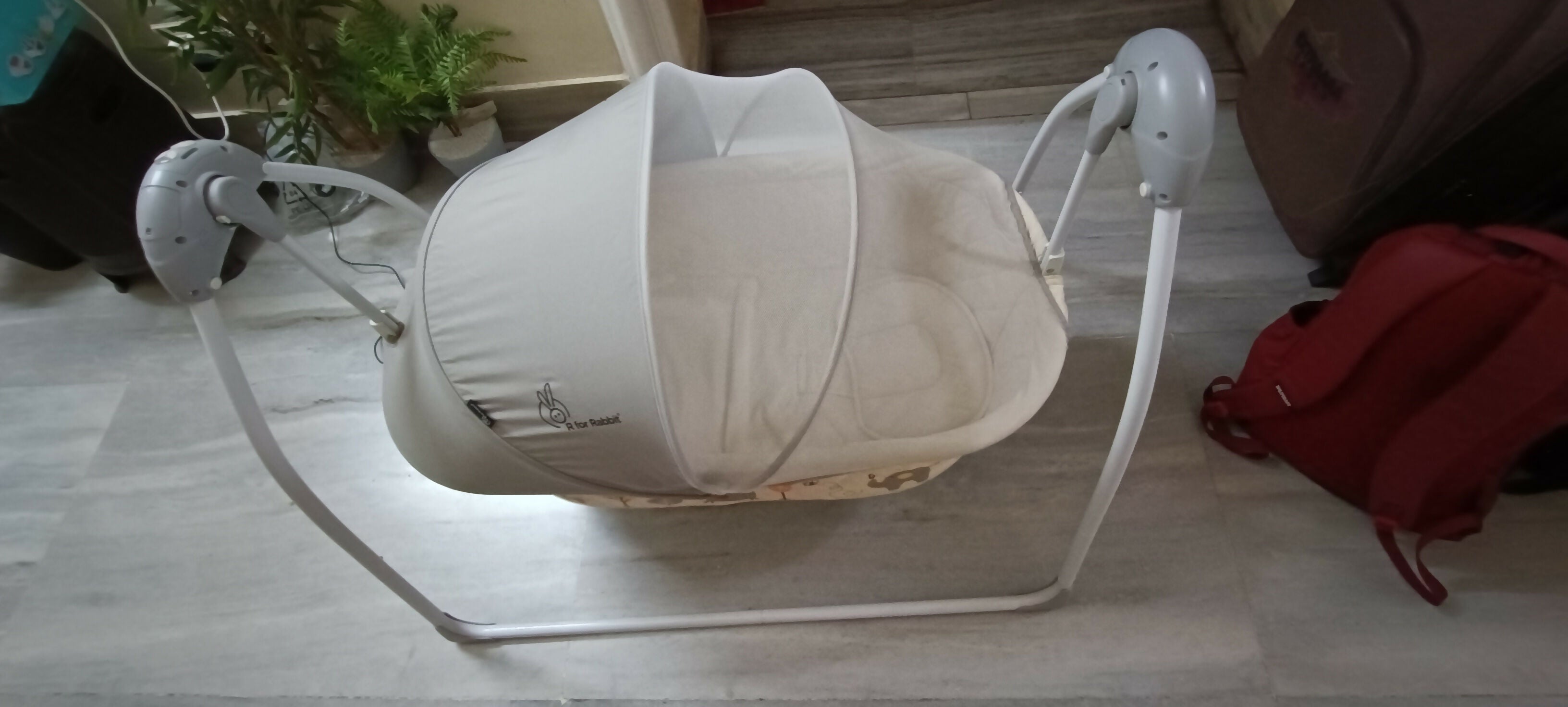 Baby swing on a wooden floor with a red bag and plant in the background