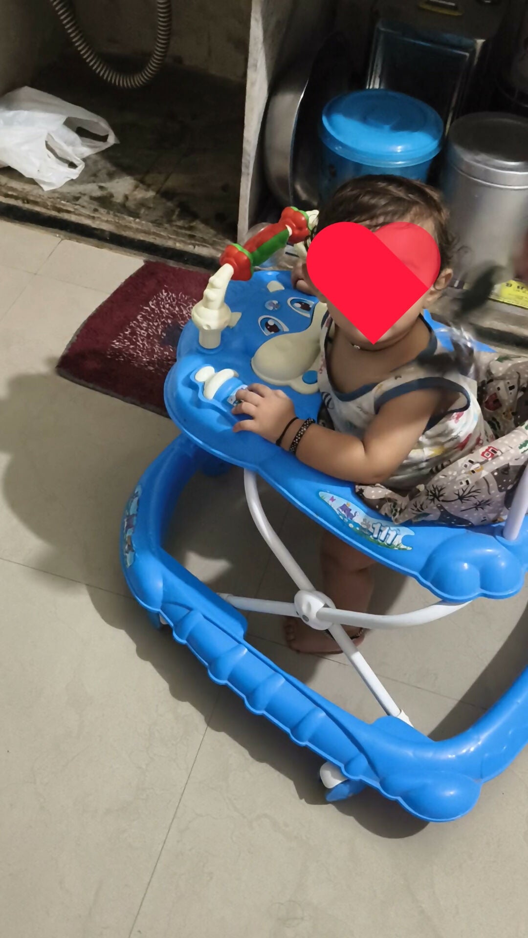 Child in a blue walker with a blurred face, sitting on a tiled floor.