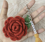 Hand holding a red crocheted flower keychain with a green leaf and yellow tasse