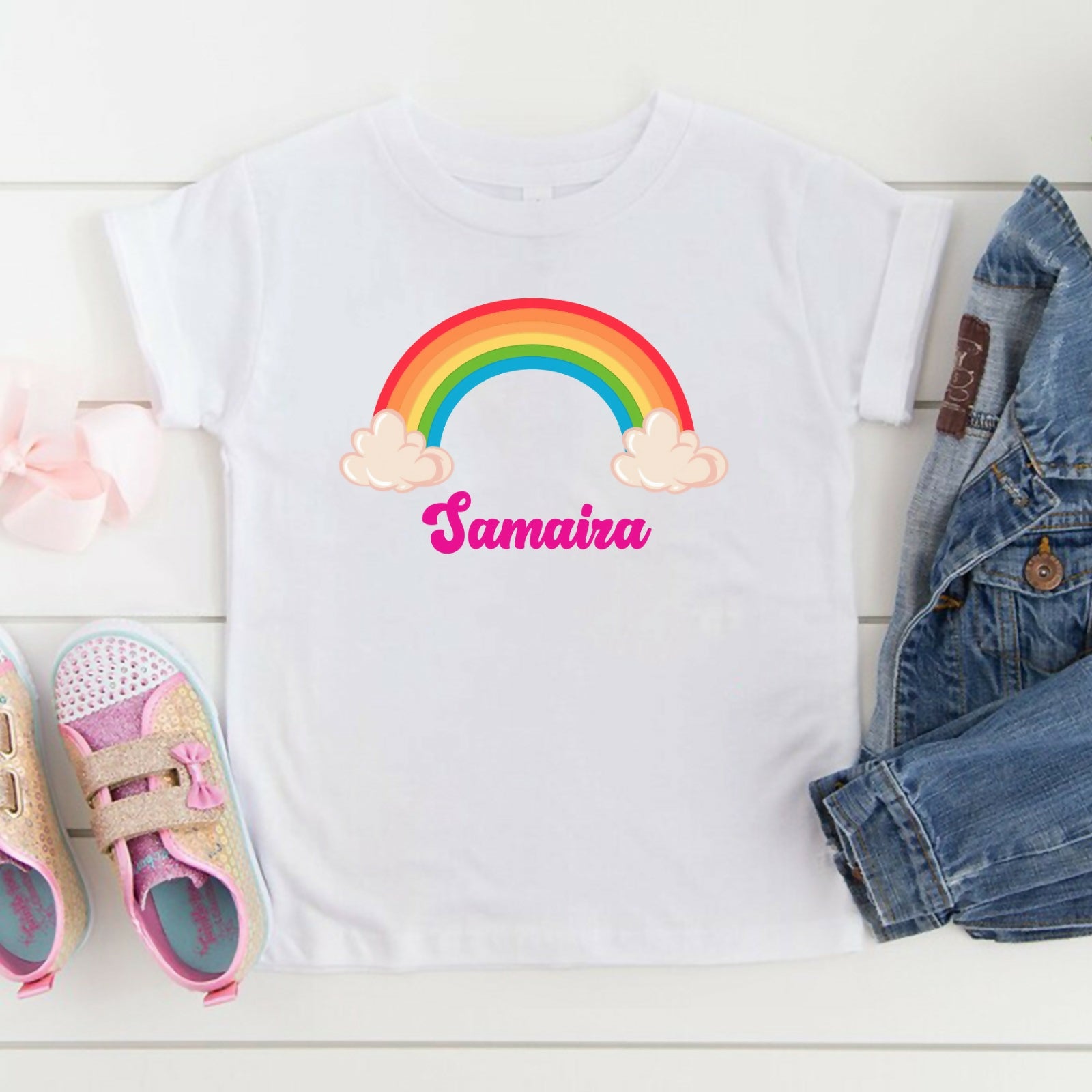 White t-shirt with rainbow and 'Samaira' design, surrounded by children's shoes and denim jacket on a white surface.