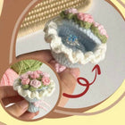 Decorative crochet flower pot with pink flowers held by a hand, on a light background.