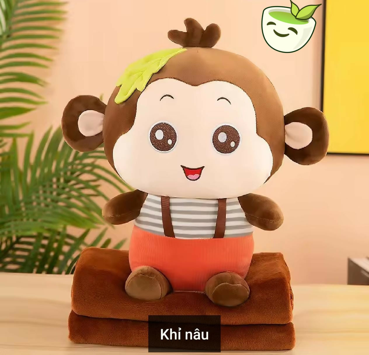 Plush monkey toy with a green leaf on its head, sitting on a brown blanket against a light background.