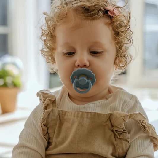 Child wearing a beige outfit with a blue pacifier indoors