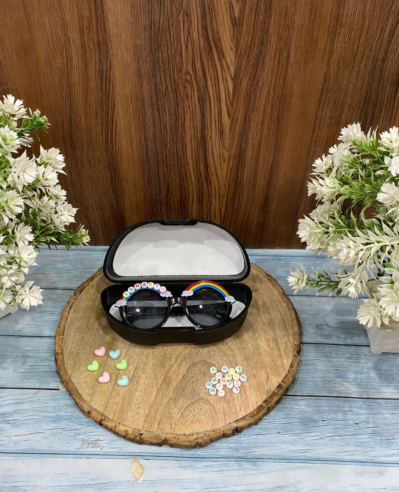 Sunglasses with decorative elements in a case on a wooden coaster with flowers in the background