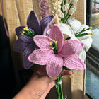Hand holding a bouquet of pink, purple, and white crocheted flowers against a neutral background.