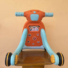 Children's toy scooter with orange seat and blue handlebars on a wooden stand against a beige wall.