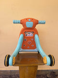 Children's toy scooter with orange seat and blue handlebars on a wooden stand against a beige wall.