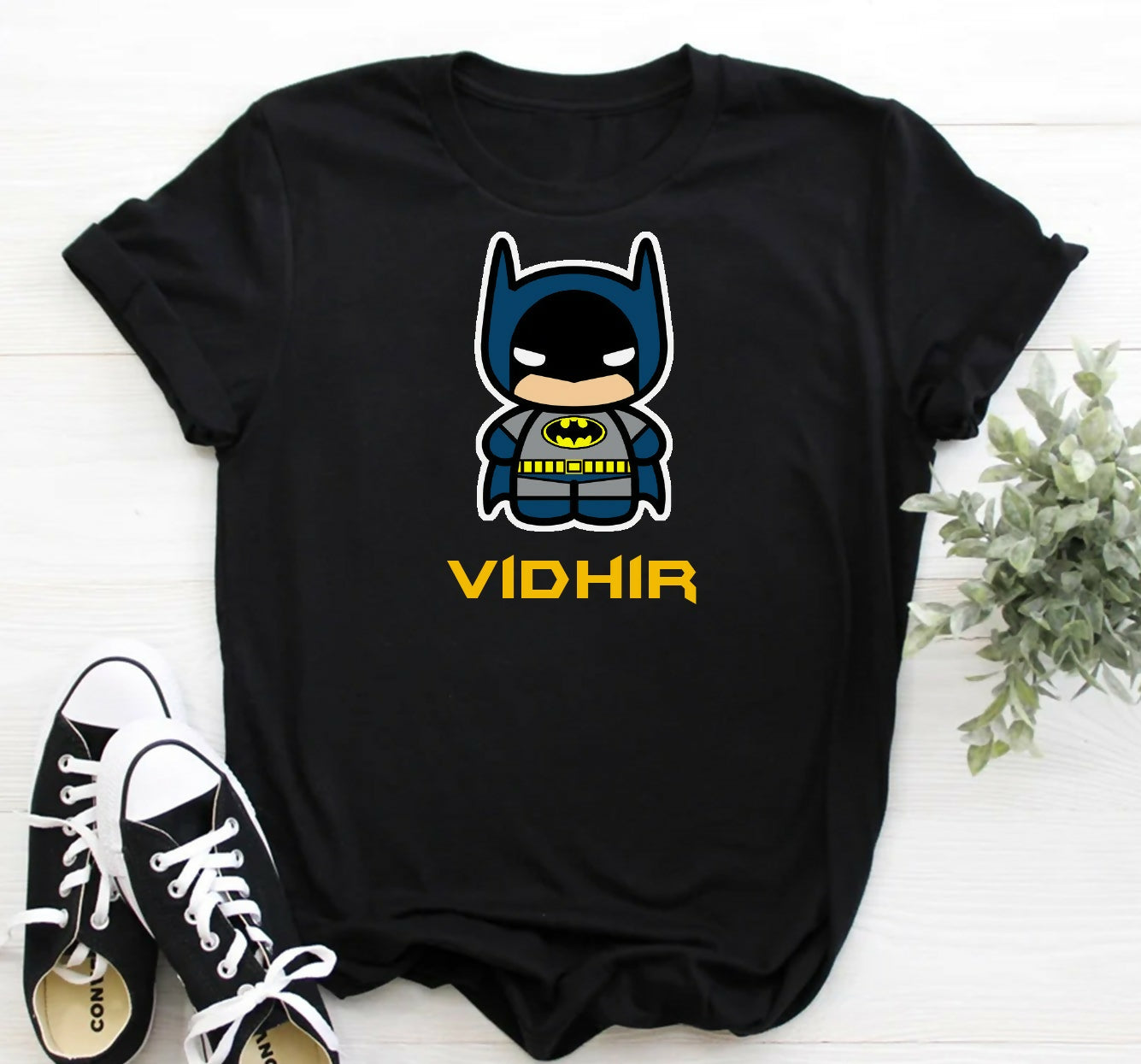 Black t-shirt with a cartoon Batman graphic and 'VIDHIR' text, paired with black and white sneakers on a light wooden surface.