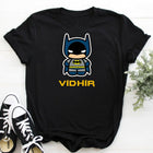 Black t-shirt with a cartoon Batman graphic and 'VIDHIR' text, paired with black and white sneakers on a light wooden surface.
