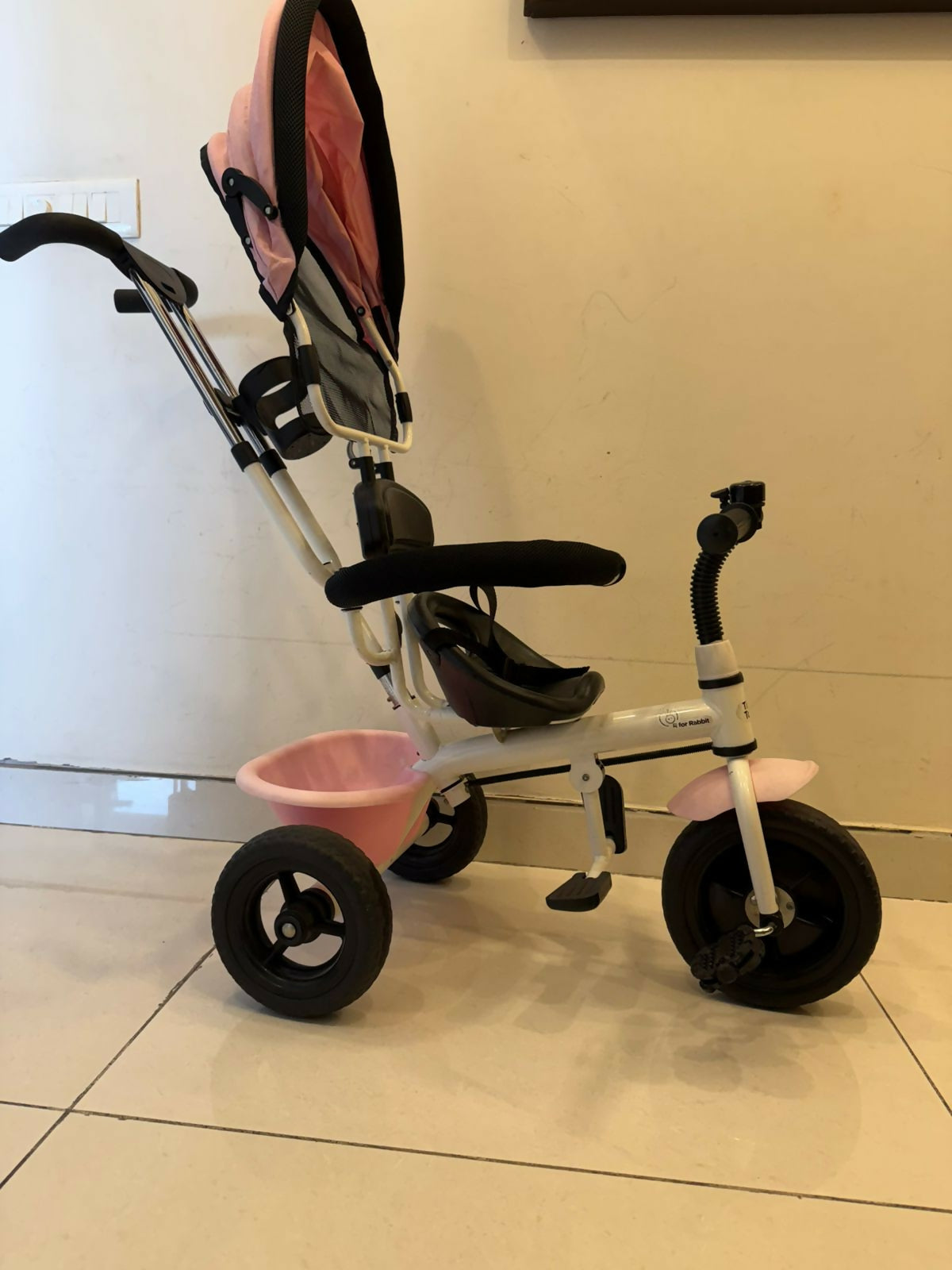 Children's tricycle with pink seat and basket on a tiled floor.