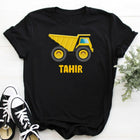 Black t-shirt with yellow dump truck graphic and 'Tahir' text, paired with black and white sneakers on a light wooden surface.