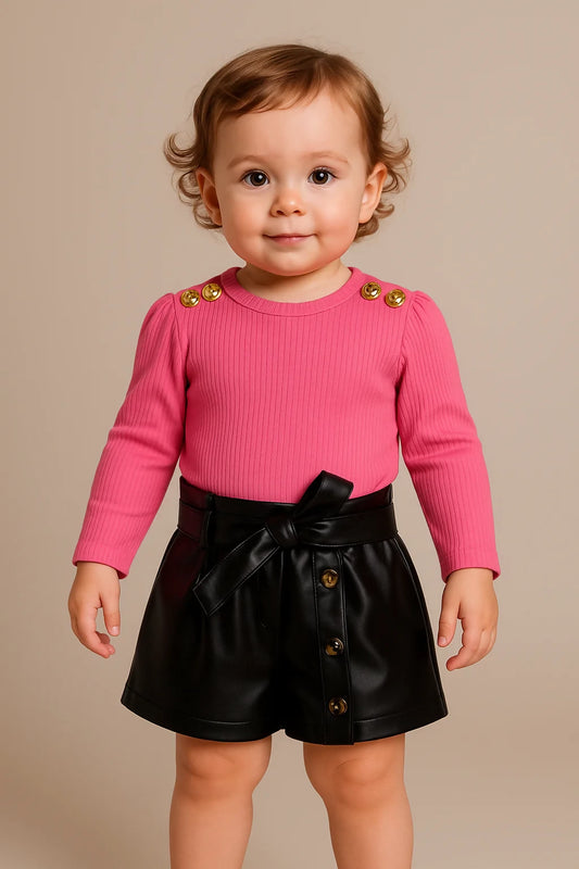 Child wearing a pink top with gold buttons and black shorts with a bow 
