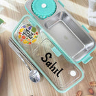 Stainless steel bento box with clear compartment, turquoise lid, and personalized name 'Sahil' on a wooden surface.