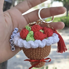 Hand holding a crocheted keychain shaped like a cupcake with red roses, green leaves, and a red bow.