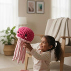 Child holding a pink knitted octopus toy in a room with a plant and chair.