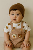 Baby wearing a brown knit hat and bear-themed overalls sitting on a white surface.
