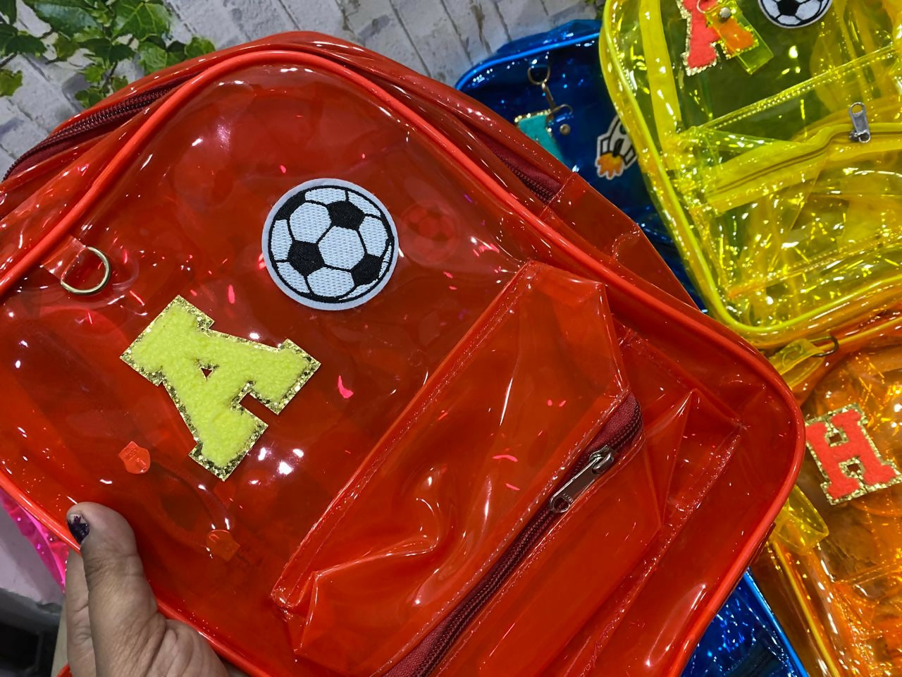 Red transparent backpack with soccer ball design and letter 'H' held by a hand, with other colorful backpacks in the background.