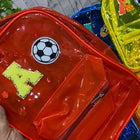 Red transparent backpack with soccer ball design and letter 'H' held by a hand, with other colorful backpacks in the background.