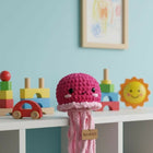 Pink crochet jellyfish toy on a shelf with colorful toys and a framed picture in the background.