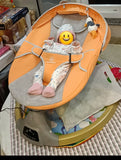 Baby in an orange and gray baby bouncer with a smiling emoji face toy.